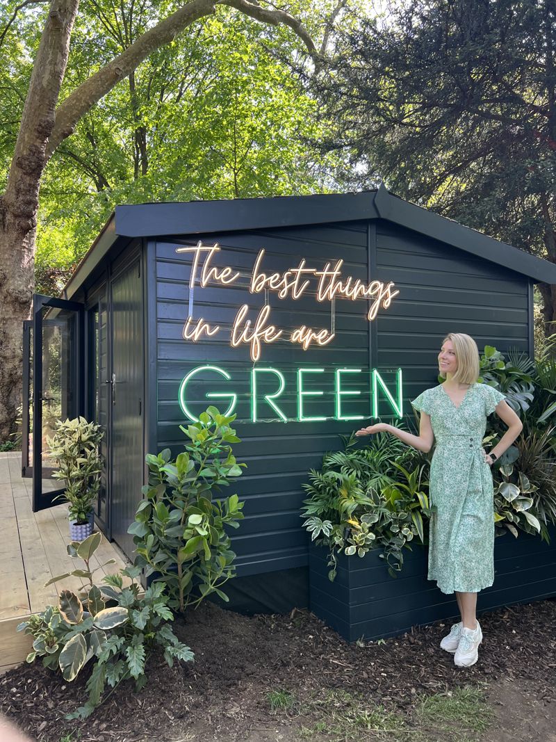 Sandy beside a dark garden studio with a neon sign reading 'The best things in life are green', surrounded by lush foliage